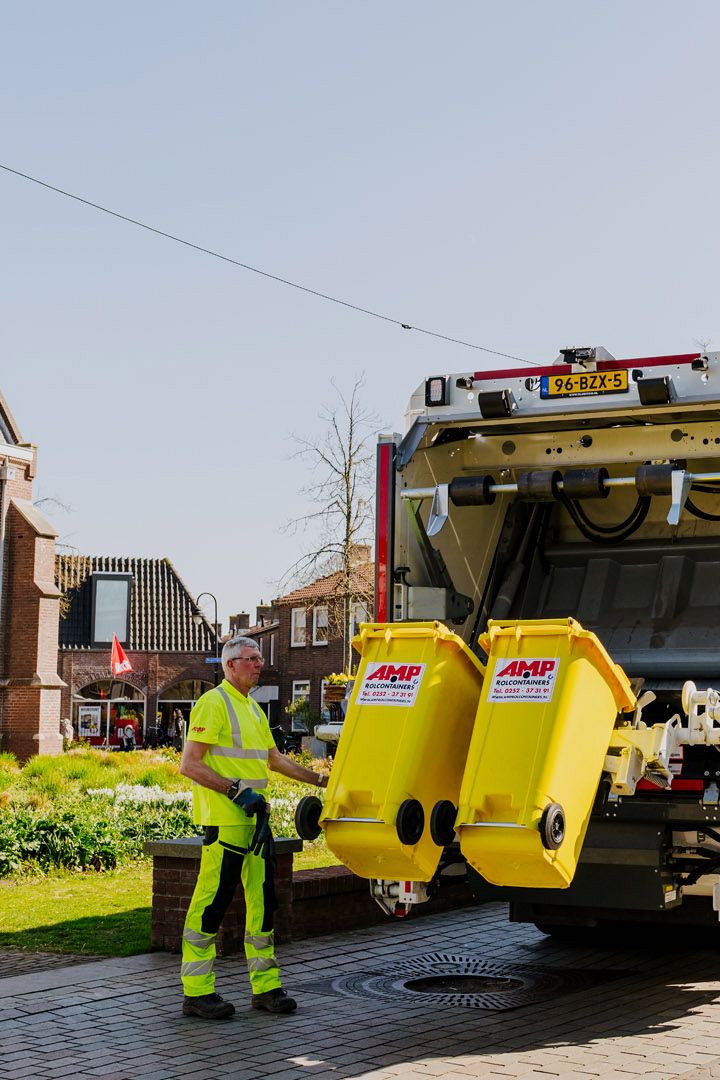 Een vuilniswagen van A.M.P. Afvalinzameling leegt gele afvalcontainers terwijl een medewerker toezicht houdt op straat.