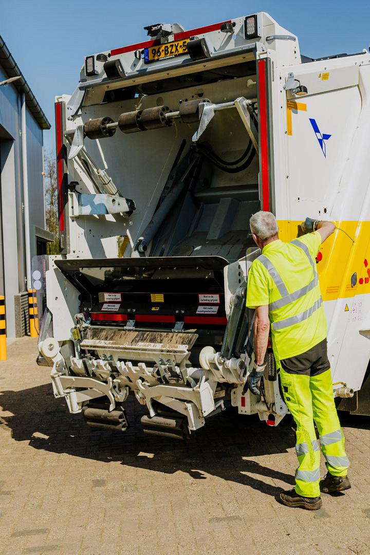 AMP-medewerker inspecteert de achterkant van een witte vuilniswagen met open laadmechanisme.