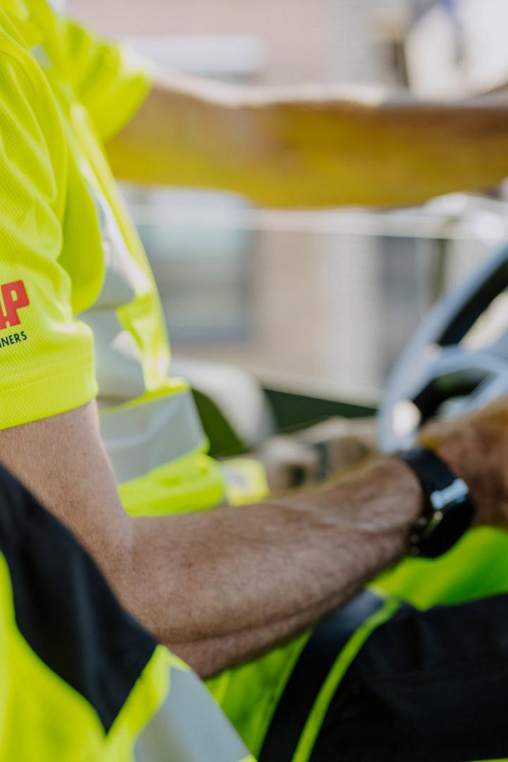 Close-up van een AMP-medewerker in fluorescerend uniform achter het stuur van een vrachtwagen.