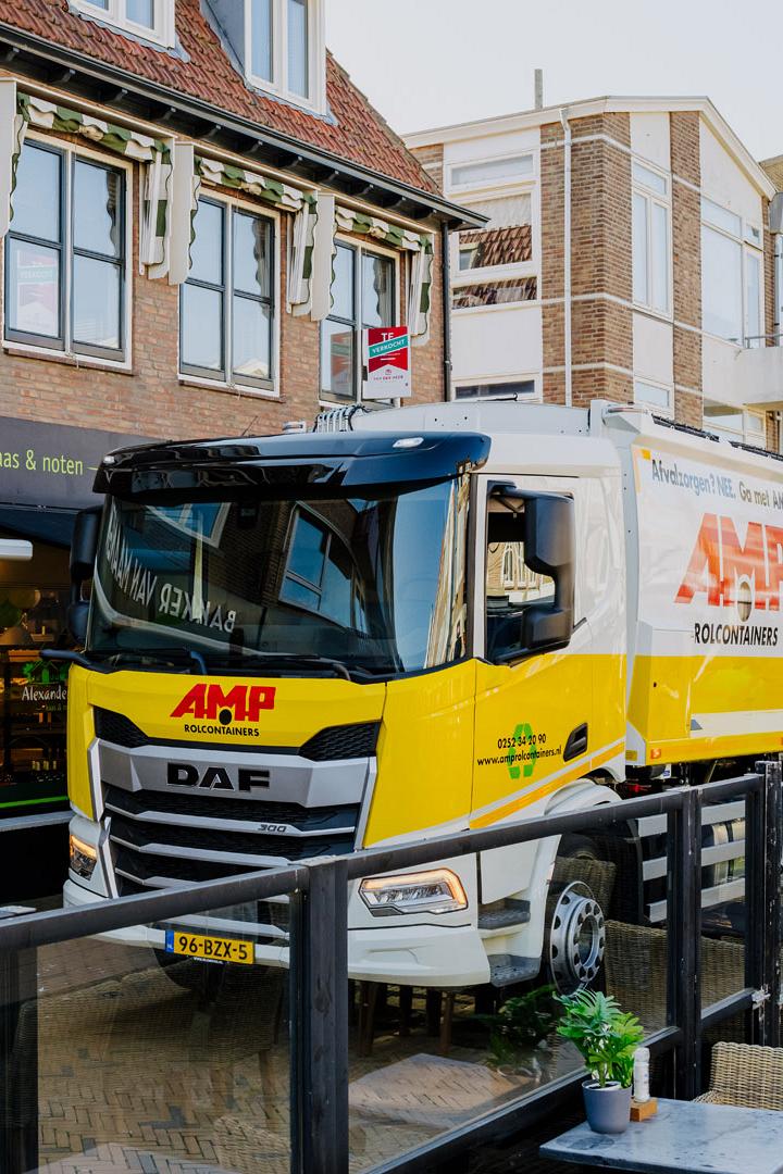 AMP-vrachtwagen manoeuvreert door een smalle winkelstraat met terras en oude gevels.
