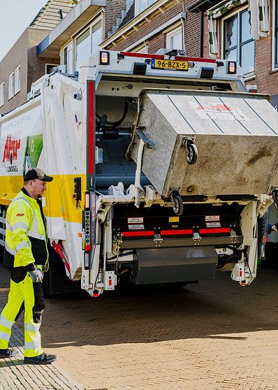 Twee medewerkers in fluorescerende kleding bedienen een vuilniswagen die een grote afvalcontainer optilt in een winkelstraat.