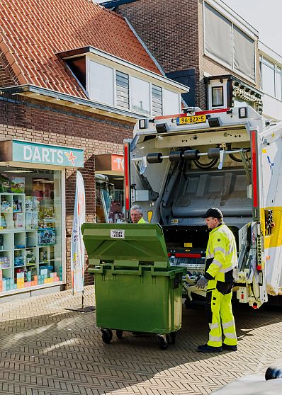 Lachende AMP-medewerker rolt een gele container langs een fietsenwinkel over de stoep.