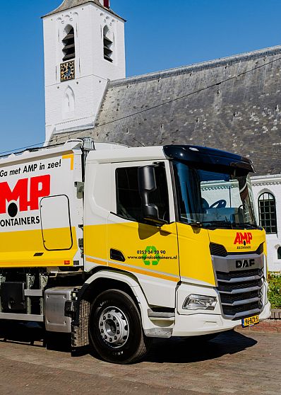 AMP-inzamelwagen staat geparkeerd bij een historische kerk, met duidelijke branding en herkenbare gele huisstijl tijdens een inzamelronde in het dorp.