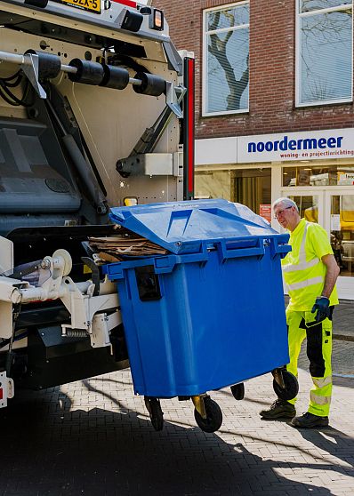 AMP-medewerker begeleidt een blauwe rolcontainer die wordt geleegd in de inzamelwagen, met zichtbaar kartonafval en duidelijk herkenbare straatomgeving.