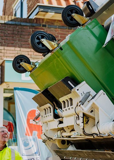 Een groene rolcontainer wordt geleegd in de AMP-inzamelwagen, met zicht op het ledigingssysteem en een medewerker die het proces volgt in een winkelstraat.