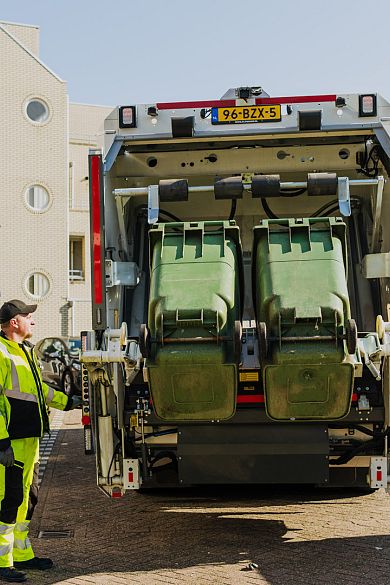Een vuilniswagen tilt twee groene afvalcontainers op in een woonstraat, terwijl een medewerker in fluorescerende werkkleding naast de wagen staat.