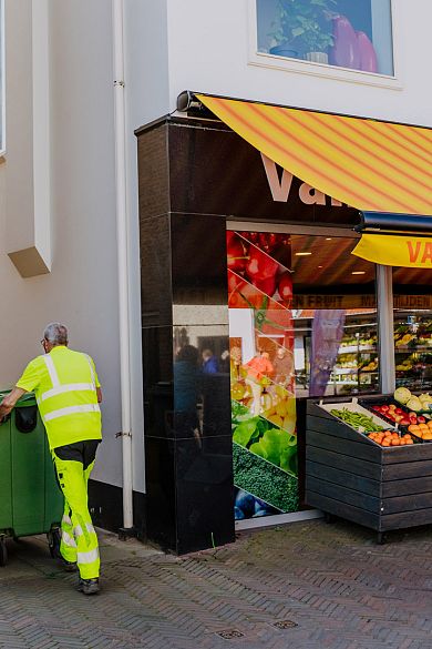 Een medewerker in fluorescerende werkkleding duwt groene afvalcontainers langs een groentewinkel met kleurrijke luifel en fruitkraam.