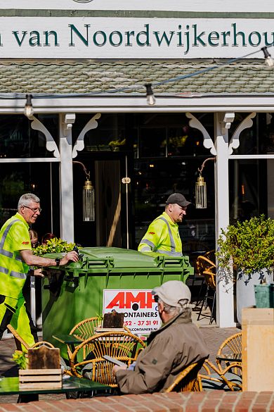 Twee medewerkers in fluorescerende kleding verplaatsen een grote groene afvalcontainer langs het terras van restaurant Het Wapen van Noordwijkerhout.