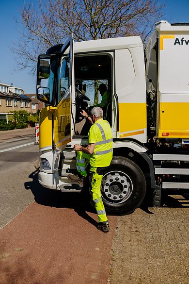 Een medewerker in fluorescerende werkkleding praat bij de open cabine van een gele en witte afvalwagen met de chauffeur.