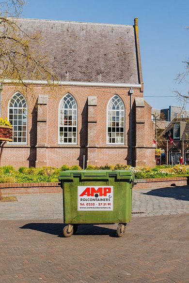 Groene AMP-rolcontainer op een plein met een bakstenen kerkgebouw op de achtergrond.