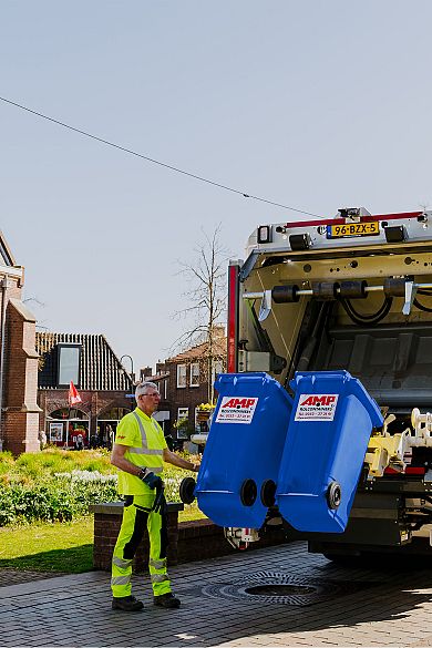 AMP-medewerker begeleidt het legen van twee blauwe containers in een vuilniswagen in een woonwijk.