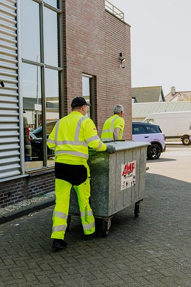 Vuilnisman leegt een grote blauwe container in een vuilniswagen voor een winkelpand.