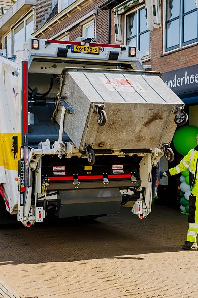 Grote metalen AMP-container wordt geleegd in een vuilniswagen in een woonwijk.