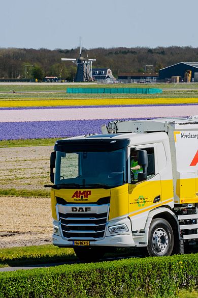 AMP-vrachtwagen rijdt door de bollenstreek met kleurrijke velden en een molen op de achtergrond.