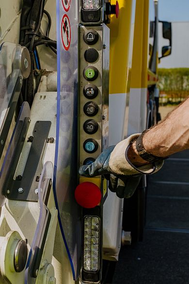 Close-up van de bedieningsknoppen op een AMP-vuilniswagen, met een medewerker die op een knop drukt.