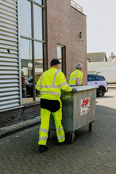 Twee AMP-medewerkers verplaatsen samen een grote metalen rolcontainer langs een bedrijfspand.