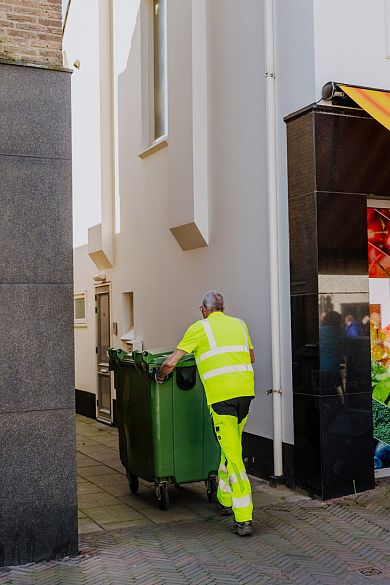 AMP-medewerker in fluorescerende werkkleding duwt een groene container door een smalle steeg naast een winkel.