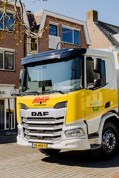 Voorzijde van een gele AMP-vrachtwagen in een woonwijk op een zonnige dag.