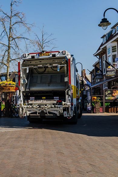 Vuilniswagen van AMP staat klaar op een plein in het stadscentrum met bloemen en winkels op de achtergrond.