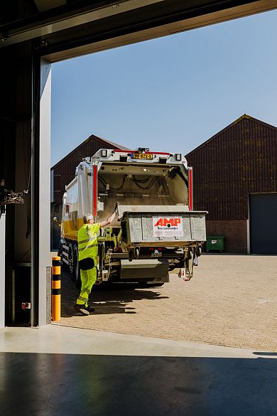 Vuilniswagen van AMP rijdt een loods uit op een bedrijventerrein, begeleid door een medewerker in veiligheidstenue.