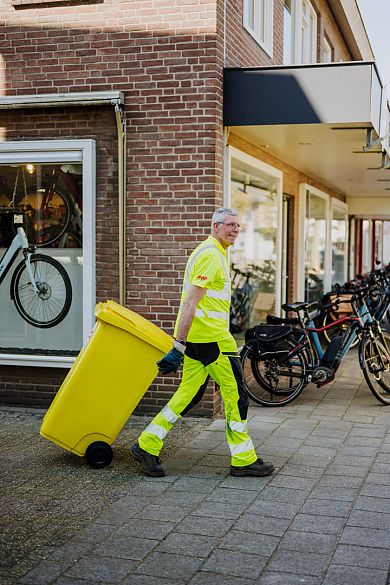 Lachende AMP-medewerker rolt een gele container langs een fietsenwinkel over de stoep.
