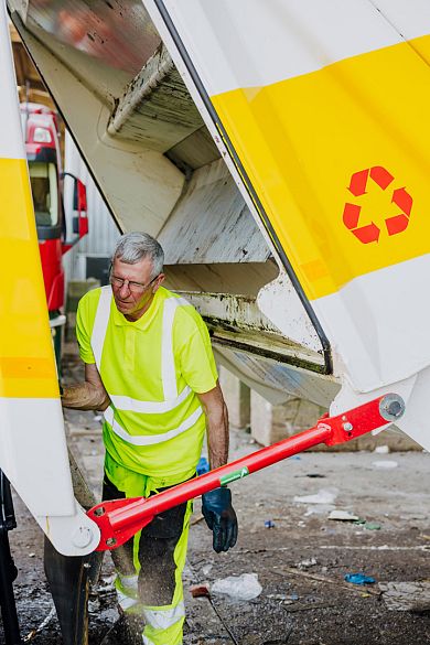 Lachende AMP-medewerker rolt een gele container langs een fietsenwinkel over de stoep.