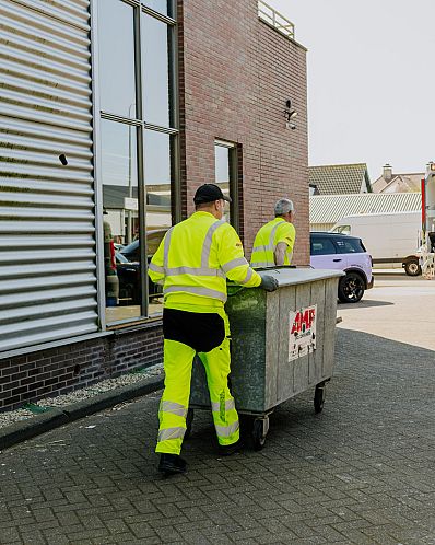 Twee AMP-medewerkers verplaatsen samen een grote metalen rolcontainer langs een bedrijfspand.