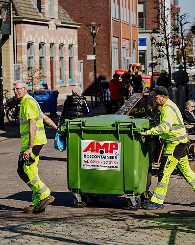 AMP-medewerker in fluorescerende werkkleding duwt een groene container door een smalle steeg naast een winkel.