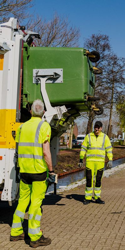 Twee AMP-medewerkers bij een vuilniswagen terwijl een grote groene container wordt geleegd.