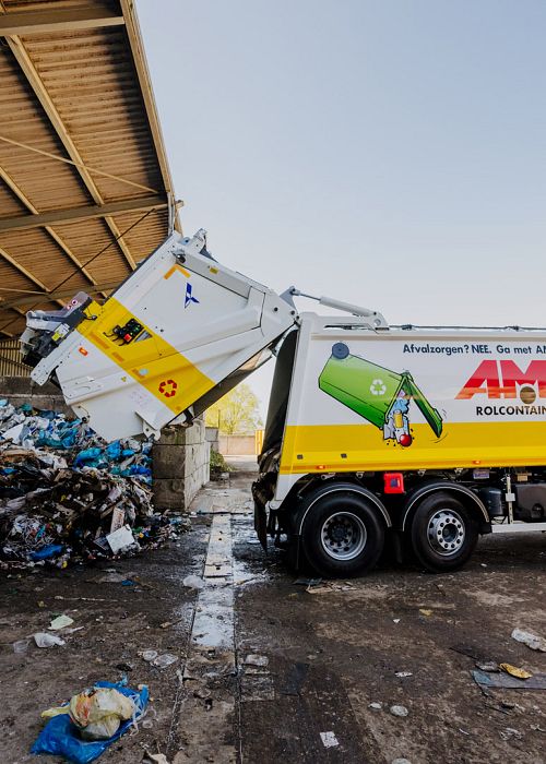 Een gele en witte vuilniswagen stort afval in een grote hoop onder een overdekte afvalverwerkingshal.