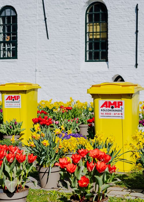 AMP containers bij witte kerk tussen de bloemen.