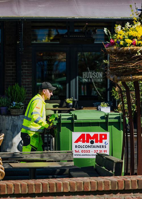  AMP-medewerker verplaatst een groene rolcontainer bij een horecagelegenheid, met duidelijk zichtbaar het AMP-logo en de containeropstelling buiten.