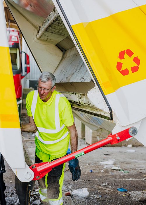 AMP-medewerker begeleidt de vuilniswagen bij het verlaten van de loods, met zicht op het achterlaadsysteem en het bedrijfsterrein op een heldere dag.