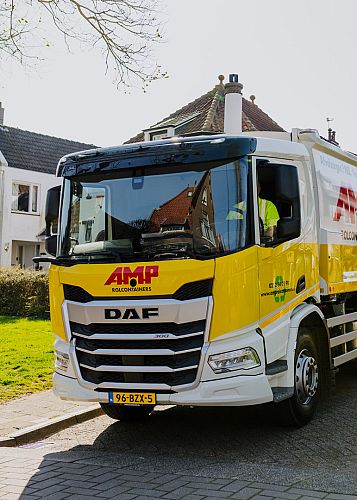 AMP-inzamelwagen rijdt door een woonwijk tijdens de inzamelronde, met duidelijke gele huisstijl en zicht op de chauffeur in de cabine.