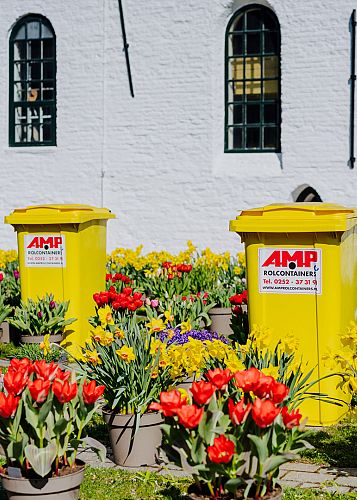 Twee gele AMP-containers staan tussen kleurrijke voorjaarsbloemen bij een wit kerkpand, als onderdeel van afvalbeheer tijdens een lokaal evenement.