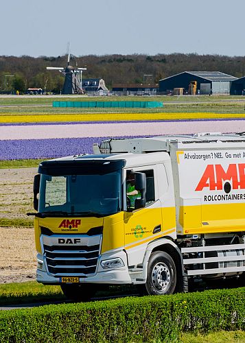AMP-inzamelwagen rijdt langs uitgestrekte bloembollenvelden, met duidelijk zichtbare branding en slogan tijdens een route door de regio.