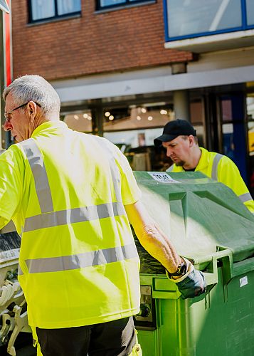 Twee medewerkers in gele werkkleding legen groene container in vuilniswagen in winkelstraat.