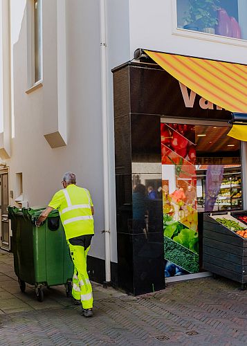 AMP-medewerker rijdt een groene rolcontainer langs een verswinkel in een drukke winkelstraat tijdens de geplande afvalinzameling.