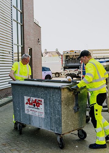 Twee medewerkers in veiligheidskleding verplaatsen een metalen rolcontainer op een bedrijventerrein richting de afvalwagen.