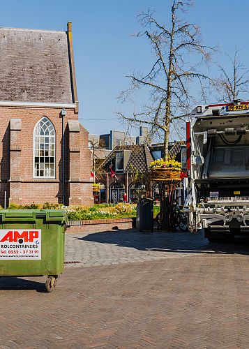 Groene AMP rolcontainer op een dorpsplein met afvalwagen op de achtergrond, klaar voor inzameling.
